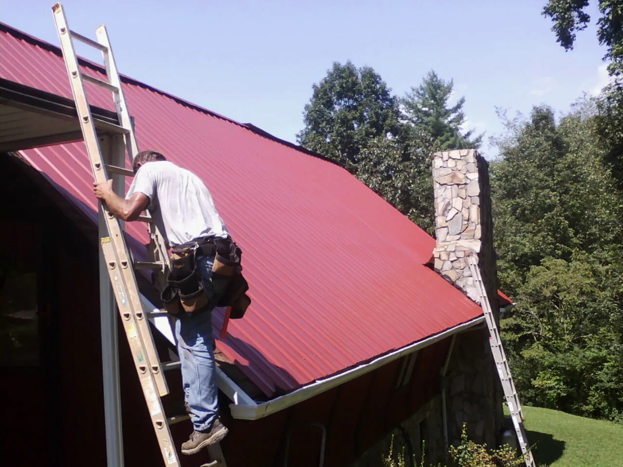 Roofer on a ladder beside a red metal roof and stone chimney — Soffit & Fascia Repair by Roofing Brothers in East Bend